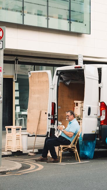 A man seated on a wooden chair beside a white moving van parked on the street in front of a modern building with large glass windows. The van's rear doors are open, revealing wrapped furniture and cardboard boxes inside, likely part of a home relocation or furniture transport process. The man is wearing glasses and a light blue shirt, preparing documents or notes, indicative of logistical planning during a difficult access removal for a basement flat in Holland Park, as part of the services provided by Man with Van Holland Park. Nearby, there are small wooden pallets and a wooden panel leaning against the van, suggesting ongoing packing or loading activities, with street markings and a corner sign visible in the background. The environment appears well-lit with natural daylight, and the setting captures the typical scene of a professional removal operation involving careful handling of packing materials and equipment for challenging access residence moves.
