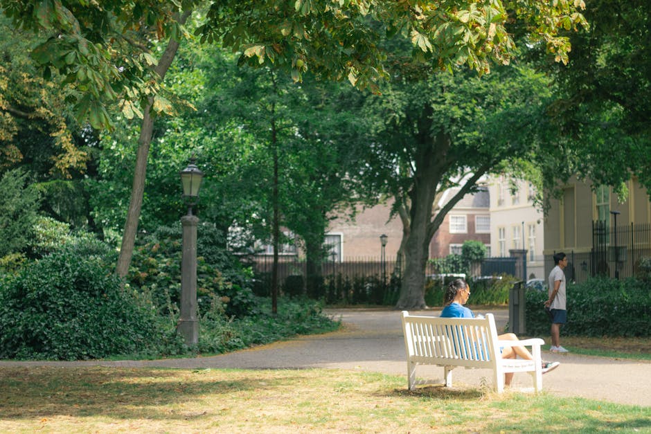 A woman with brown hair tied back, wearing a blue top and white shorts, is seated on a white wooden park bench. She is looking away from the camera, toward a young boy standing nearby, who is dressed in a white shirt and dark shorts. The scene is set in a green, leafy park with tall trees providing shade, and a paved walking path visible in the background. The park features a lamppost next to the woman and is enclosed by a black metal fence, with some residential buildings partially visible beyond the trees. The sunlight filters through the dense foliage, casting dappled shadows on the ground. This image captures a casual moment outdoors, relevant to house removals or relocation services by Man with Van Holland Park, with an emphasis on transporting items through challenging access areas such as basement flats, highlighting the importance of careful packing and logistical planning during home relocation or furniture transport.