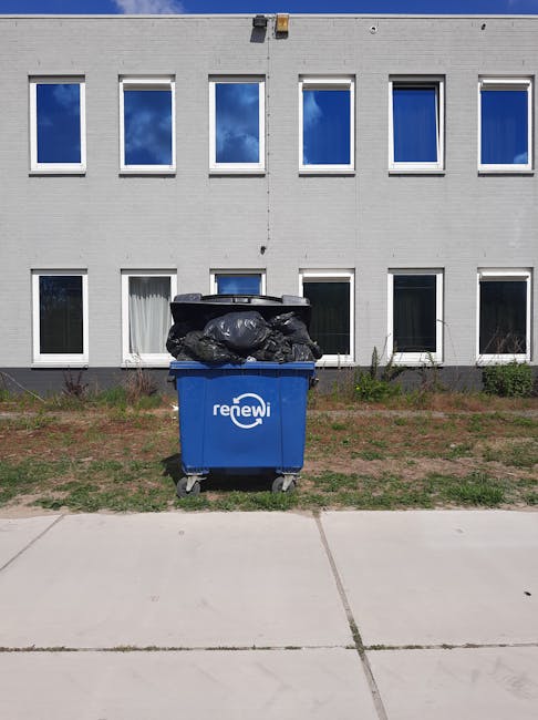 A large white and dark blue garbage collection truck parked on a paved street in front of a modern multi-storey brick building with several windows. The truck features a closed main container for wast