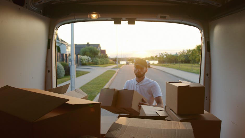 A man wearing a light-colored polo shirt and cap is seen outside the rear of a moving van during a home relocation process, standing on a residential street at sunset. Inside the van, there are several cardboard boxes, some with flaps open and others sealed, arranged on the van’s floor near the doorway. The boxes vary in size and are packed in preparation for transportation, with some stacked and others awaiting loading or unloading. The surrounding environment includes a sidewalk, lawns, trees, and residential houses, with the sunset casting a warm glow over the scene. This image illustrates the loading process involved in furniture transport and packing for house removals, with visible packaging materials and the outdoor setting typical of professional removal services such as those offered by Man with Van Holland Park.