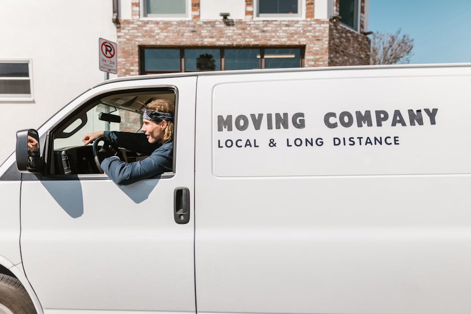 A side view photograph of a white moving van parked on a street outside a residential building with brick exterior walls and large windows. The van features the company's branding with the text 'MOVING COMPANY' and 'LOCAL & LONG DISTANCE' printed on the side. Inside the open driver’s cabin, a person wearing a dark jacket and a headband is seated, adjusting or inspecting items on the dashboard. The vehicle is positioned near a sidewalk with a no-parking sign visible nearby. The scene captures the loading process inside the property with visible furniture, boxes wrapped in plastic or cardboard, and packing materials possibly being prepared or loaded for a house relocation, illustrating the logistics involved in furniture transport and packing and moving services by Man with Van Holland Park.