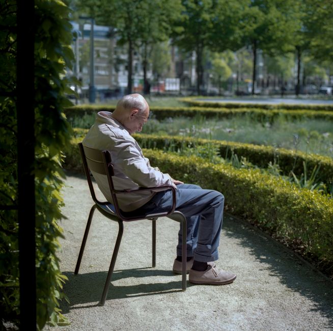 A senior man with light-colored hair sitting on a black metal and wooden chair outdoors in a park-like setting with neatly trimmed hedges and lush green trees in the background. He is wearing a light-colored jacket, blue trousers, and grey shoes, with his head bowed and eyes closed, appearing to be resting or in a moment of reflection. The ground is paved, and the area is well-lit by natural daylight, suggesting a calm and peaceful environment. This scene may relate to house removals or moving logistics, reflecting a break during a packing or transportation process. Man with Van Holland Park often provides services for home relocation, furniture transport, and packing and moving, which aligns with the serene setting shown in the image.