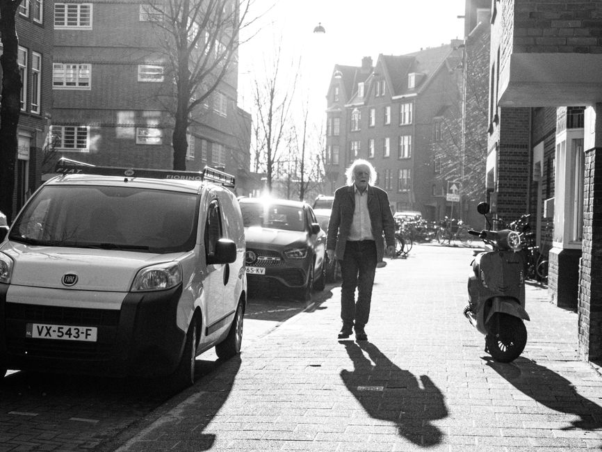 A man with white hair, dressed in a dark jacket and light-colored shirt, is walking along a residential pavement near a row of parked vehicles, including a white Fiat van with the license plate VX-543-F and a dark car behind it. The scene is set in Holland Park, Kensington and Chelsea, with multi-storey brick and masonry buildings lining the street, some with bay windows and pitched roofs. To the right of the man, a motorcycle or scooter is parked next to a brick building, with a bicycle rack further down the sidewalk. The pavement is made of brick or stone, and there are bicycles visible in the background, along with some leafless trees and a bright light source casting long shadows. This scene illustrates the logistics of home relocation and furniture transport, consistent with professional removals services offered by Man with Van Holland Park, especially in the context of obtaining moving permits for Holland Park's residential areas.