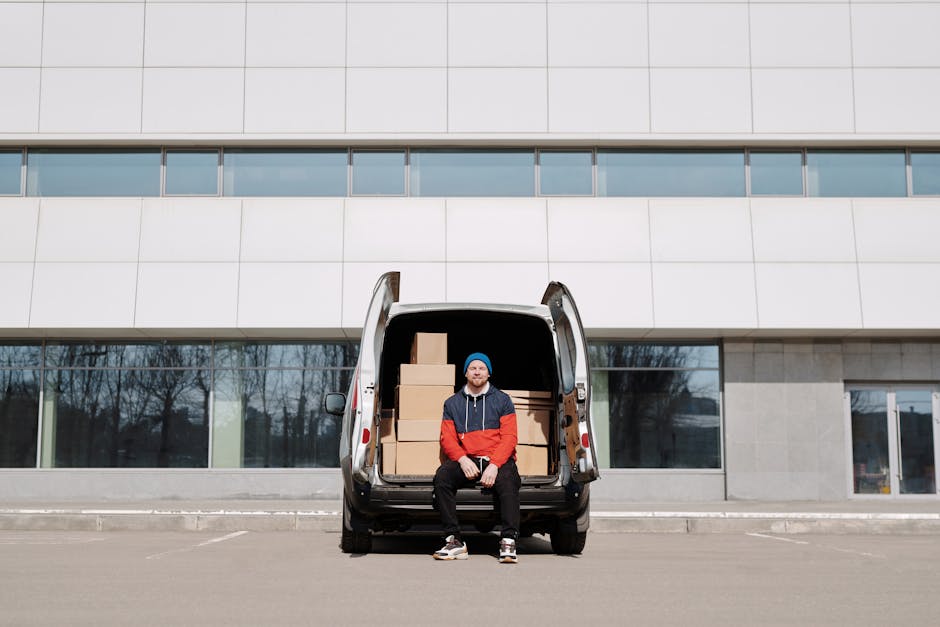 A man wearing a blue beanie, a red and navy jacket, and black pants is sitting on the open tailgate of a black van parked outside a modern building with large windows and white panels. Behind him are several packed cardboard boxes stacked inside the vehicle, ready for a home relocation or furniture transport. The van is positioned on a paved area, possibly a driveway or loading zone, and the background includes the building’s exterior and reflections in the windows. The scene depicts the loading process in a professional removals setting, with items prepared for delivery or move, and the environment is well-lit with natural daylight. Man with Van Holland Park, a company specialising in removals, is visible in this image, illustrating a typical loading and packing stage of their same-day house moving services.