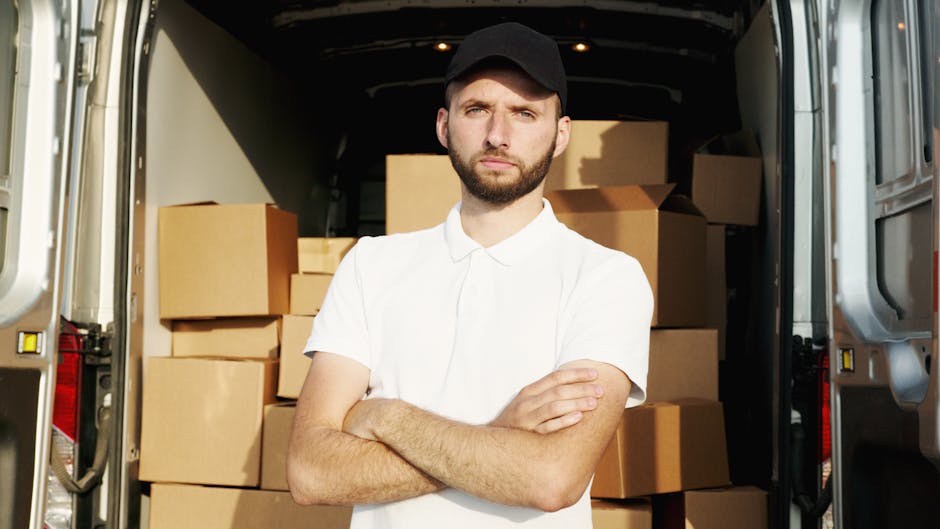 A young man with a serious expression, wearing a black cap and white polo shirt, stands inside a moving van with open rear doors. Behind him, there are numerous cardboard boxes of various sizes, some stacked on top of each other, indicating packing activities related to house removals. The interior of the van is lined with dark material, and the boxes are sealed with packing tape, suggesting careful preparation for a home relocation or furniture transport. The van is parked on a pavement, with natural daylight illuminating the scene, and the man appears to be part of a professional removals service such as Man with Van Holland Park, preparing for a relocation or loading process.