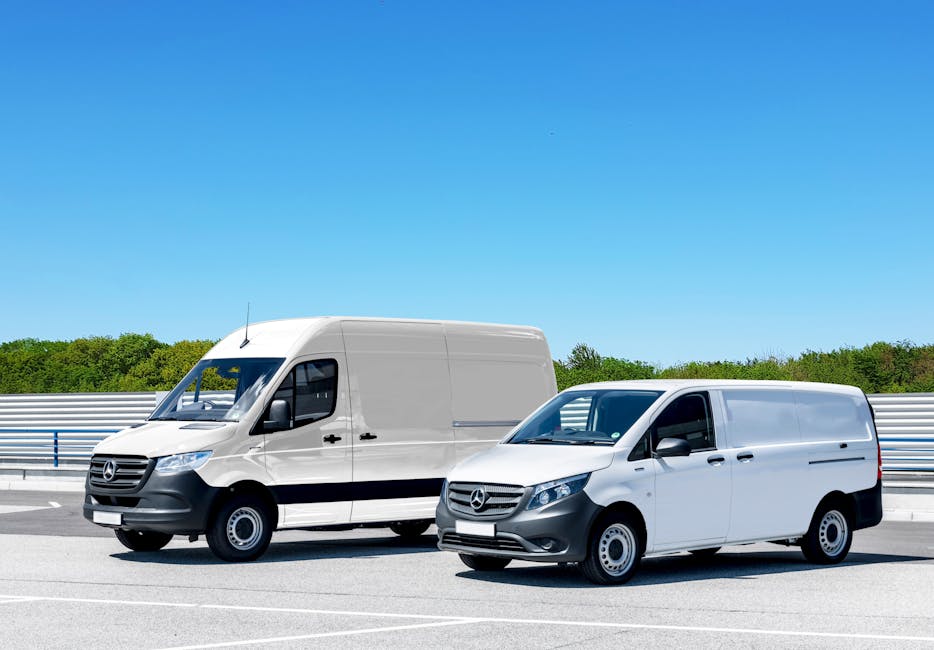 Two white Mercedes-Benz vans parked side by side on a paved surface near Holland Park station, with a metal barrier and green trees in the background under a clear blue sky. The larger van on the left is positioned slightly behind the smaller van on the right, with the doors and windows visible. The vehicles are part of a home relocation or furniture transport operation carried out by Man with Van Holland Park, as seen in the context of loading or moving processes. The scene highlights professional transport equipment, such as the vans themselves, and suggests an organized move involving packing, loading, and transit within an urban environment.