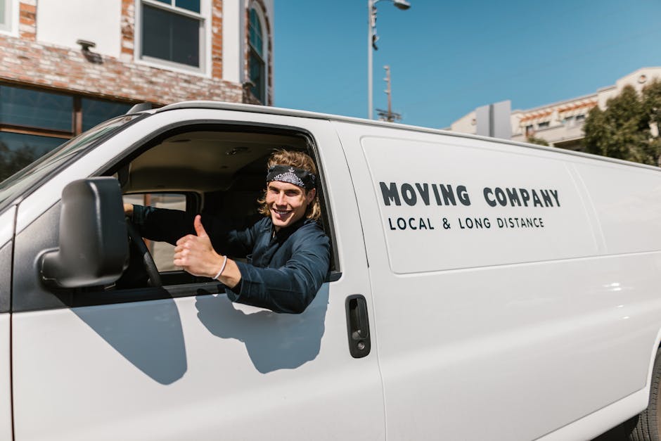 A young male mover with shoulder-length hair wearing a black and white patterned headband and a dark blue shirt sits in the driver's seat of a white moving van labeled 'MOVING COMPANY LOCAL & LONG DISTANCE.' He is smiling and giving a thumbs-up gesture through the open driver’s window, indicating readiness for home relocation or furniture transport. The van is parked outside a residential building on a sunny day, with a clear blue sky visible. Inside the vehicle, parts of packing materials, such as cardboard boxes and moving blankets, can be seen in the background, suggesting preparation for household moving or packing and moving services. The scene captures the loading process or vehicle preparation typical of professional removals carried out by Man with Van Holland Park, reflecting organized transport and relocation logistics.