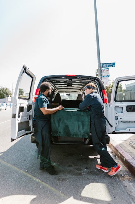 Two movers from Man with Van Holland Park are loading a large, dark green upholstered piece of furniture into the open rear of a white van parked on a street. The interior of the van reveals a spacious cargo area with a black and silver interior. One mover, on the left, wears a dark t-shirt and trousers, while the other, on the right, is dressed in a dark jacket, light trousers, and red shoes, both engaged in lifting and guiding the furniture into the vehicle. The scene takes place outdoors on a bright day, with a street sign visible above on a lamppost and some buildings or shops in the background, indicating a typical urban setting suitable for home relocation or furniture transport. The image captures the physical effort involved in the loading process during a house removal, highlighting the careful handling of belongings and professional approach of the service offered by Man with Van Holland Park.