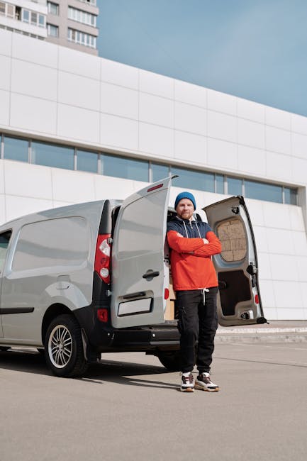 A man wearing a blue beanie and red jacket stands with arms crossed beside an open grey cargo van parked on a paved driveway in front of a modern white building with large windows and curved architectural lines. Inside the van's cargo area, visible cardboard boxes and packing materials are arranged, indicating preparation for a house removal or moving process. The van is positioned close to the building's entrance, which is likely used for loading and unloading furniture and boxes during home relocation. Natural daylight illuminates the scene, highlighting the orderly setup for furniture transport and packing and moving activities, with no other objects or people visible in the vicinity. This image reflects the logistical aspects of house removals and the professional approach taken by Man with Van Holland Park.