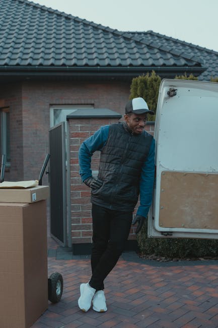 Two movers from Man with Van Holland Park are loading a large, dark green upholstered piece of furniture into the open rear of a white van parked on a street. The interior of the van reveals a spacious cargo area with a black and silver interior. One mover, on the left, wears a dark t-shirt and trousers, while the other, on the right, is dressed in a dark jacket, light trousers, and red shoes, both engaged in lifting and guiding the furniture into the vehicle. The scene takes place outdoors on a bright day, with a street sign visible above on a lamppost and some buildings or shops in the background, indicating a typical urban setting suitable for home relocation or furniture transport. The image captures the physical effort involved in the loading process during a house removal, highlighting the careful handling of belongings and professional approach of the service offered by Man with Van Holland Park.